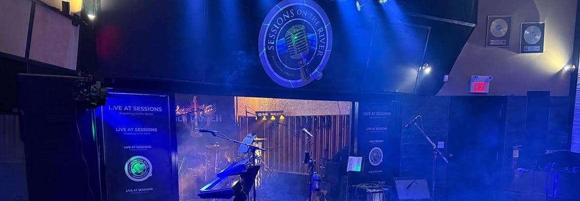 The Sessions on the River live room lit blue, with the venue's microphone badge above the stage and a drum kit and keyboard set up below